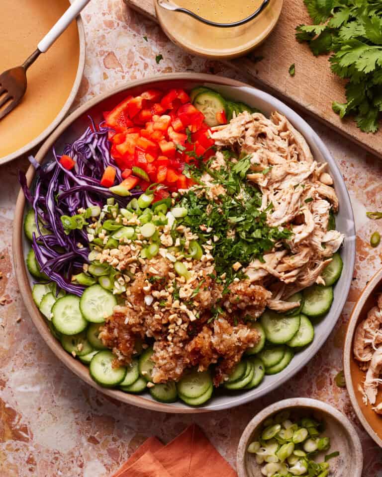 A colorful salad with shredded chicken, sliced cucumber, red bell pepper, purple cabbage, green onions, and chopped peanuts, garnished with cilantro. A bowl of dressing and a cutting board with cilantro are in the background.