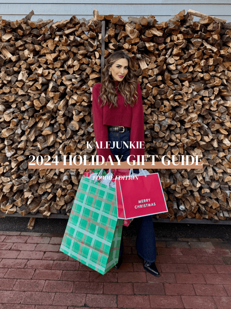 A woman in a red sweater and jeans poses in front of a large stack of firewood, holding two gift bags. The image has text that reads "Kalejunkie 2024 Holiday Gift Guide" and "Foodie Edition.