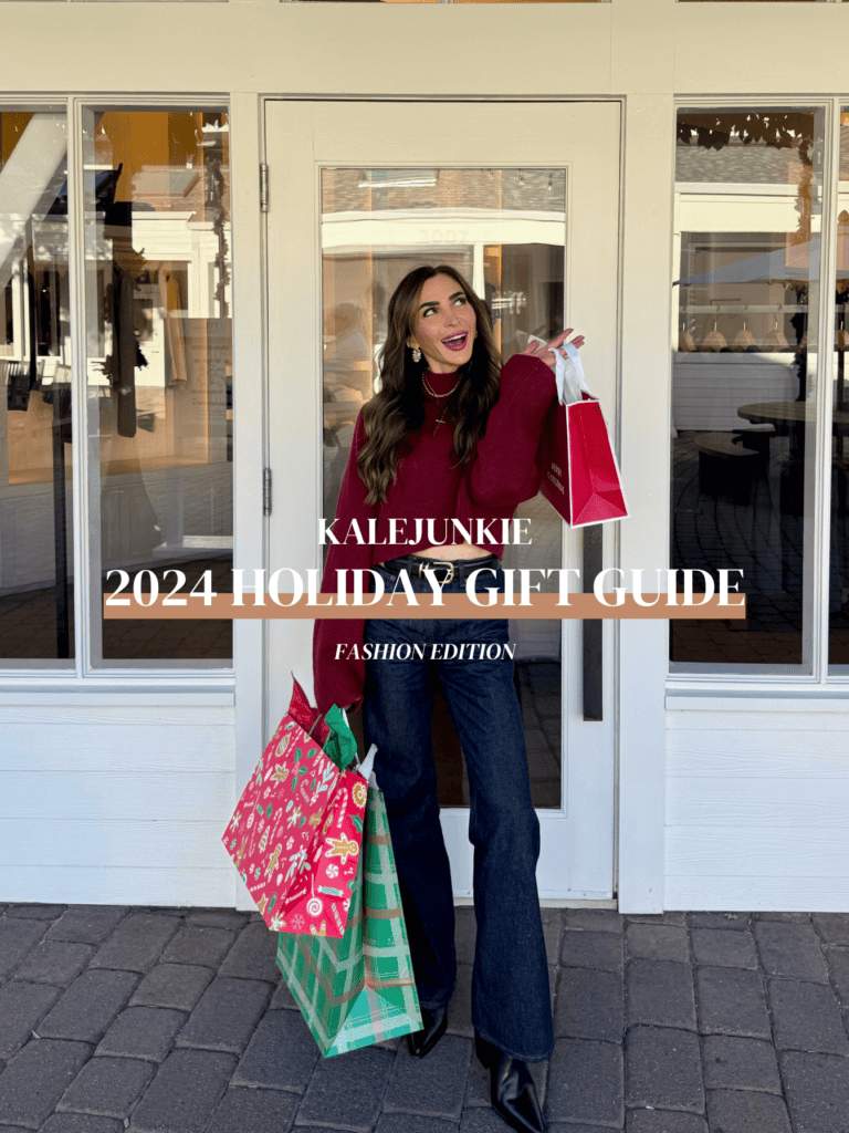 A woman in a burgundy sweater holds colorful shopping bags and smiles in front of glass doors. Overlaid text reads "Kalejunkie 2024 Holiday Gift Guide Fashion Edition." The setting is a shopping area with a festive vibe.