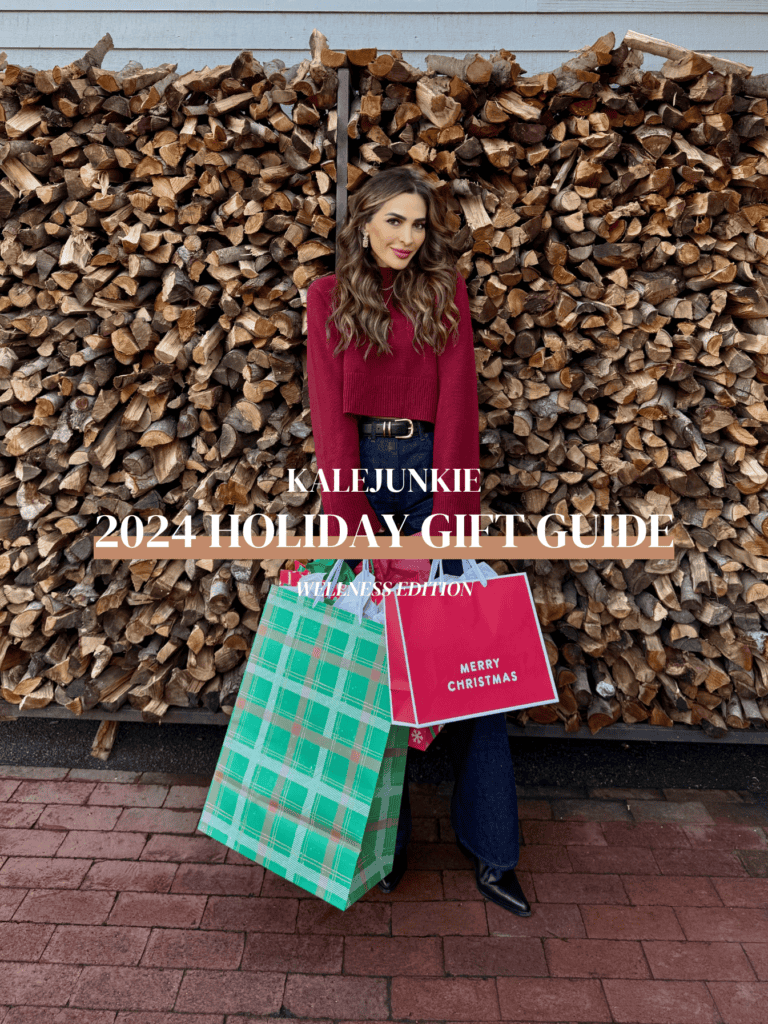 A person stands in front of a stacked woodpile, holding two large gift bags. The bags are green plaid and red with "Merry Christmas" written on them. The image is labeled "Kalejunkie 2024 Holiday Gift Guide Wellness Edition.