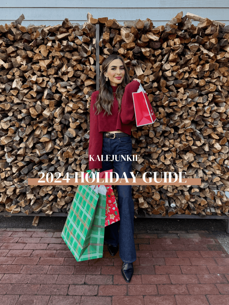 A woman in a red sweater and jeans stands in front of a large stack of firewood, holding a red gift bag. She is also holding green and red shopping bags. Text on the image reads "KALEJUNKIE 2024 HOLIDAY GUIDE.