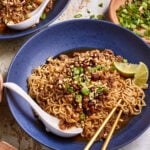 A blue bowl filled with noodle soup topped with minced meat, chopped green onions, and red chili flakes. A lime wedge sits on the side. Chopsticks rest on the bowl's edge, with a spoon nearby. Another bowl and chopped green onions are in the background.