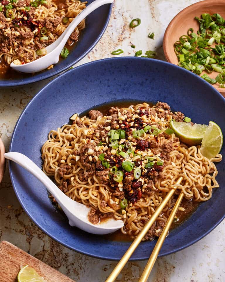A blue bowl filled with noodle soup topped with minced meat, chopped green onions, and red chili flakes. A lime wedge sits on the side. Chopsticks rest on the bowl's edge, with a spoon nearby. Another bowl and chopped green onions are in the background.