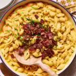 A skillet filled with creamy mac and cheese topped with crispy bacon pieces and chopped chives. A wooden spoon rests in the dish, ready to serve. Plates and a cloth napkin are seen in the background.