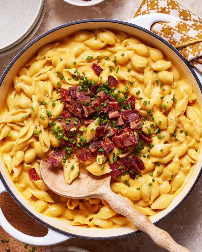 A skillet filled with creamy mac and cheese topped with crispy bacon pieces and chopped chives. A wooden spoon rests in the dish, ready to serve. Plates and a cloth napkin are seen in the background.