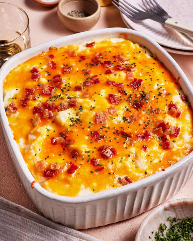 A baked casserole topped with melted cheese, bacon bits, and herbs in a white dish. Nearby are a fork, knife, glass of wine, and small bowls of seasoning. The table is set with a light pink tablecloth.