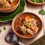 Two bowls of beef and barley soup filled with chunks of beef, vegetables, and barley are set on a table. Fresh parsley and a ladle are nearby, along with a green napkin. The scene conveys a warm, hearty meal ready to be enjoyed.