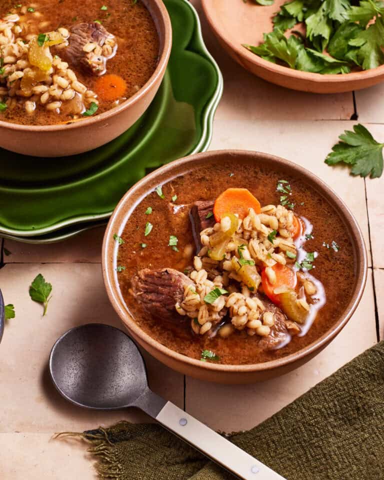 Two bowls of beef and barley soup filled with chunks of beef, vegetables, and barley are set on a table. Fresh parsley and a ladle are nearby, along with a green napkin. The scene conveys a warm, hearty meal ready to be enjoyed.