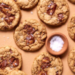 Chocolate chip cookies with chunks of chocolate and a sprinkle of salt are arranged on a peach-colored background. A small bowl of coarse sea salt is nestled among the cookies.