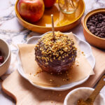 A caramel apple covered in chocolate and chopped nuts on a parchment-lined dish. Surrounding it are apples, a bowl of chocolate chips, and a small bowl of chopped nuts on a marble surface.