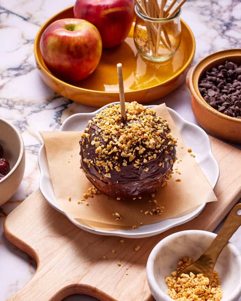 A caramel apple covered in chocolate and chopped nuts on a parchment-lined dish. Surrounding it are apples, a bowl of chocolate chips, and a small bowl of chopped nuts on a marble surface.