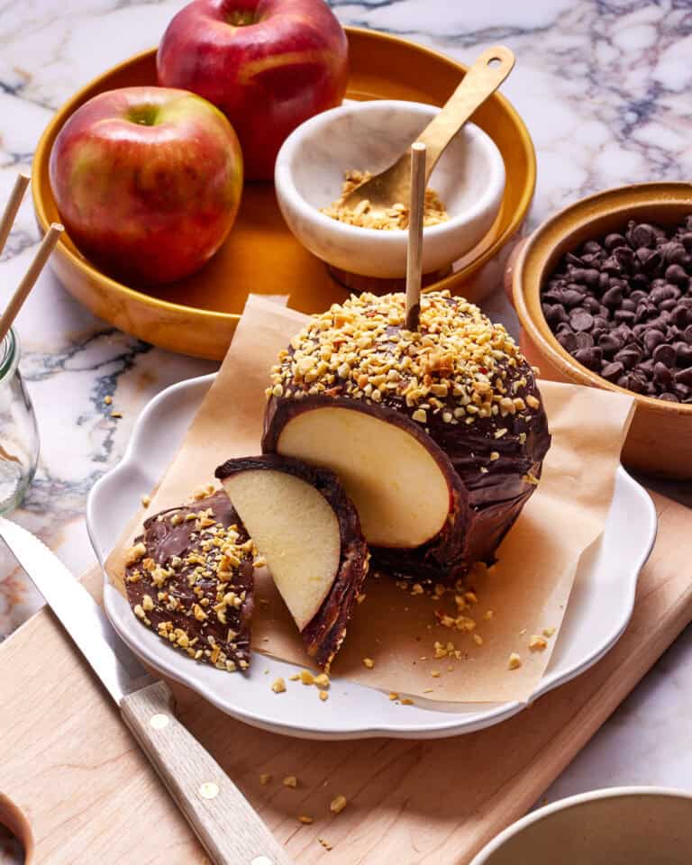 A chocolate-coated apple, topped with chopped nuts, is sliced on a parchment-covered plate. Nearby are whole apples, a bowl of chocolate chips, and a small dish containing more nuts on a marble surface. A knife rests on a wooden board.