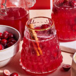 A glass of red fruit punch garnished with a cinnamon stick sits on a table. Nearby are a bowl of fresh cranberries, a glass pitcher with more punch, and a few scattered cranberries. The scene is set against a warm, red backdrop.