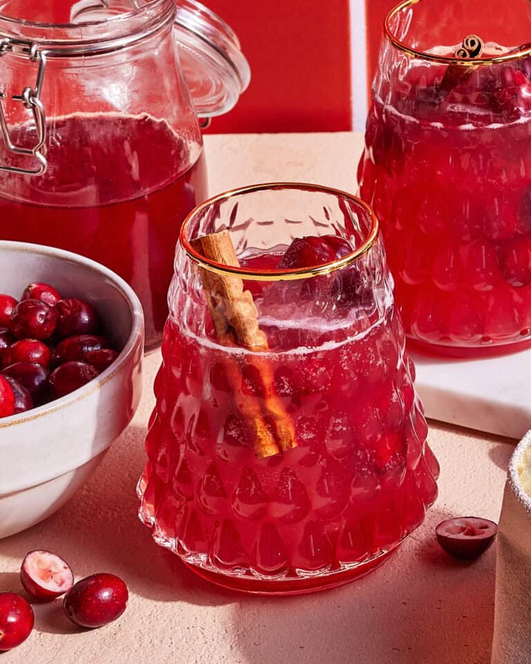 A glass of red fruit punch garnished with a cinnamon stick sits on a table. Nearby are a bowl of fresh cranberries, a glass pitcher with more punch, and a few scattered cranberries. The scene is set against a warm, red backdrop.
