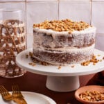 A two-layer naked cake with white frosting and a generous sprinkle of chopped nuts on top sits on a white cake stand. Surrounding items include a wicker glass, two gold forks, an empty plate, and a small wooden bowl with more nuts.