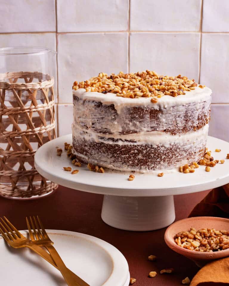 A two-layer naked cake with white frosting and a generous sprinkle of chopped nuts on top sits on a white cake stand. Surrounding items include a wicker glass, two gold forks, an empty plate, and a small wooden bowl with more nuts.