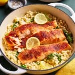 A white pot filled with cooked salmon filets on a bed of pearled couscous, spinach, and sliced lemons. A smaller bowl with couscous is in the background, alongside whole lemons and a small dish of pepper. A yellow cloth is nearby.