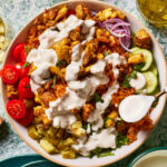 A bowl filled with shawarma-style chicken, sliced cherry tomatoes, cucumber, pickles, red onion, and diced herbs, all drizzled with a creamy white sauce. A spoon rests on the bowl's rim, and a small dish of olive oil is nearby.