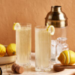 Two tall glasses filled with a light, bubbly drink garnished with lemon slices. The setting includes lemons, a wooden tray, a cork, a wire cage, a shaker, and ice cubes on a marble surface against a peach-colored wall.