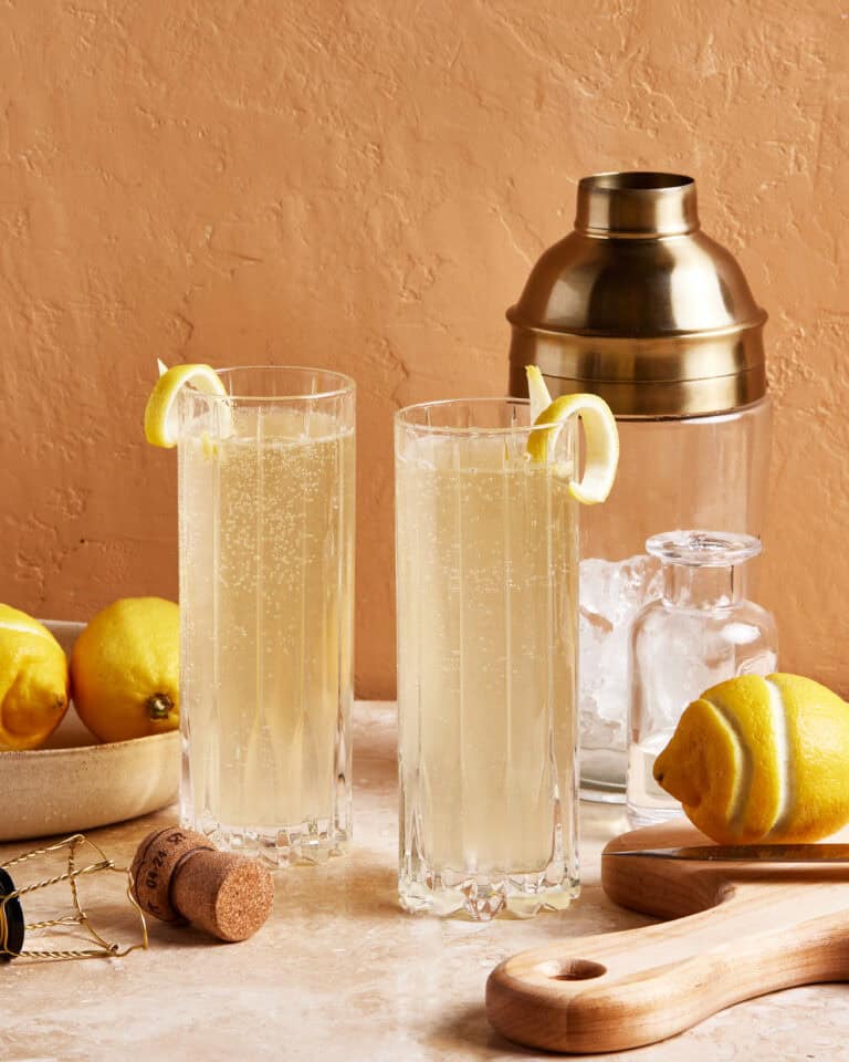 Two tall glasses filled with a light, bubbly drink garnished with lemon slices. The setting includes lemons, a wooden tray, a cork, a wire cage, a shaker, and ice cubes on a marble surface against a peach-colored wall.