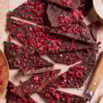 Broken pieces of dark chocolate bark topped with vibrant red freeze-dried raspberries are scattered on parchment paper. A knife rests nearby, and powdered sugar lightly dusts some pieces, adding contrast and texture.