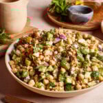 A vibrant and fresh salad in a bowl featuring chickpeas, diced cucumbers, red onions, fresh herbs, and crumbled feta cheese. A jug and fresh herbs are visible in the background, giving a homely kitchen vibe.