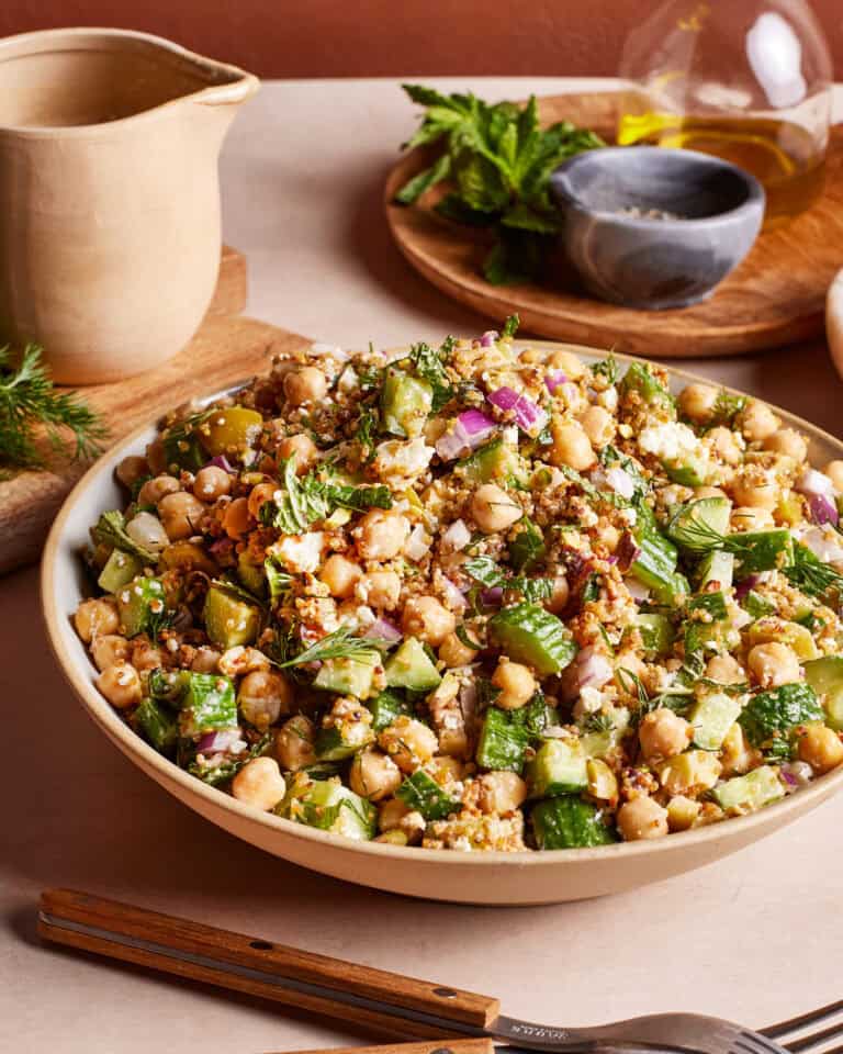 A vibrant and fresh salad in a bowl featuring chickpeas, diced cucumbers, red onions, fresh herbs, and crumbled feta cheese. A jug and fresh herbs are visible in the background, giving a homely kitchen vibe.