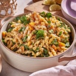A beige bowl filled with gemelli pasta, mixed with kale, breadcrumbs, and cheese. The dish is garnished with parsley. In the background, lemon halves and a wooden cutting board are visible. A pink napkin is partially seen on the right side.