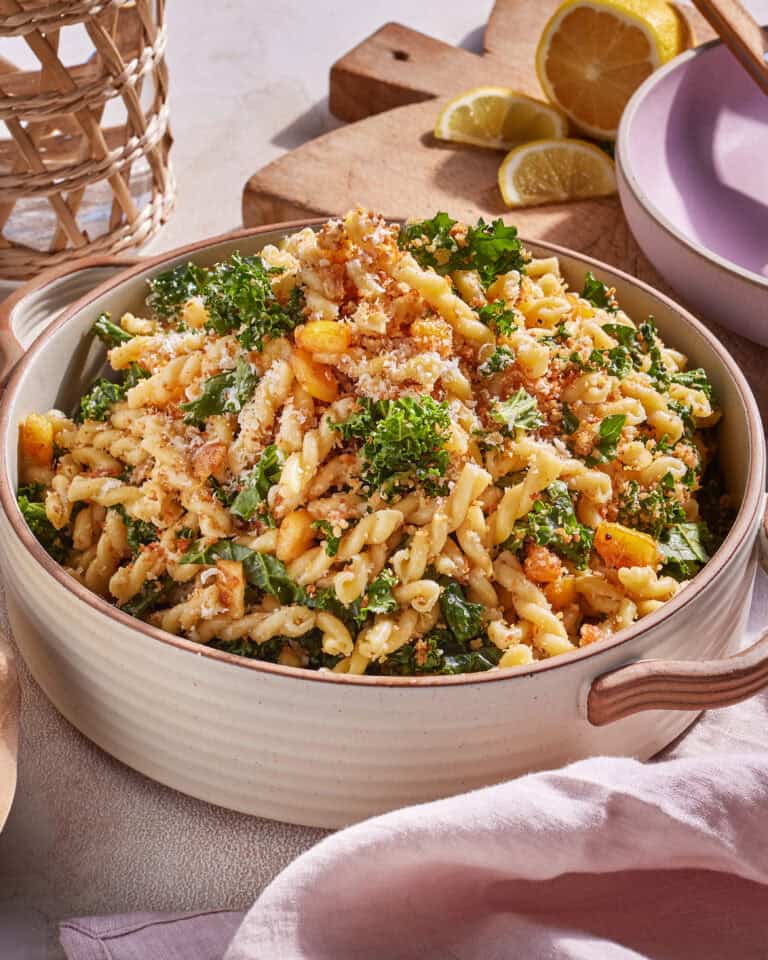 A beige bowl filled with gemelli pasta, mixed with kale, breadcrumbs, and cheese. The dish is garnished with parsley. In the background, lemon halves and a wooden cutting board are visible. A pink napkin is partially seen on the right side.