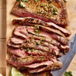 Sliced grilled beef steak garnished with chopped herbs and jalapeños on a wooden cutting board, accompanied by lime wedges and cilantro sprigs. A knife lies beside the steak.