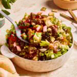 A bowl of fresh, vibrant salad with chopped lettuce, cherry tomatoes, radishes, cucumbers, and crispy pieces of toasted bread. A large spoon rests in the salad. Mint leaves and a bowl of croutons are in the background on the table.