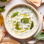 A bowl of tzatziki sauce garnished with fresh mint leaves and olive oil, surrounded by pieces of pita bread, whole mint sprigs, and two metal spoons on a green napkin.