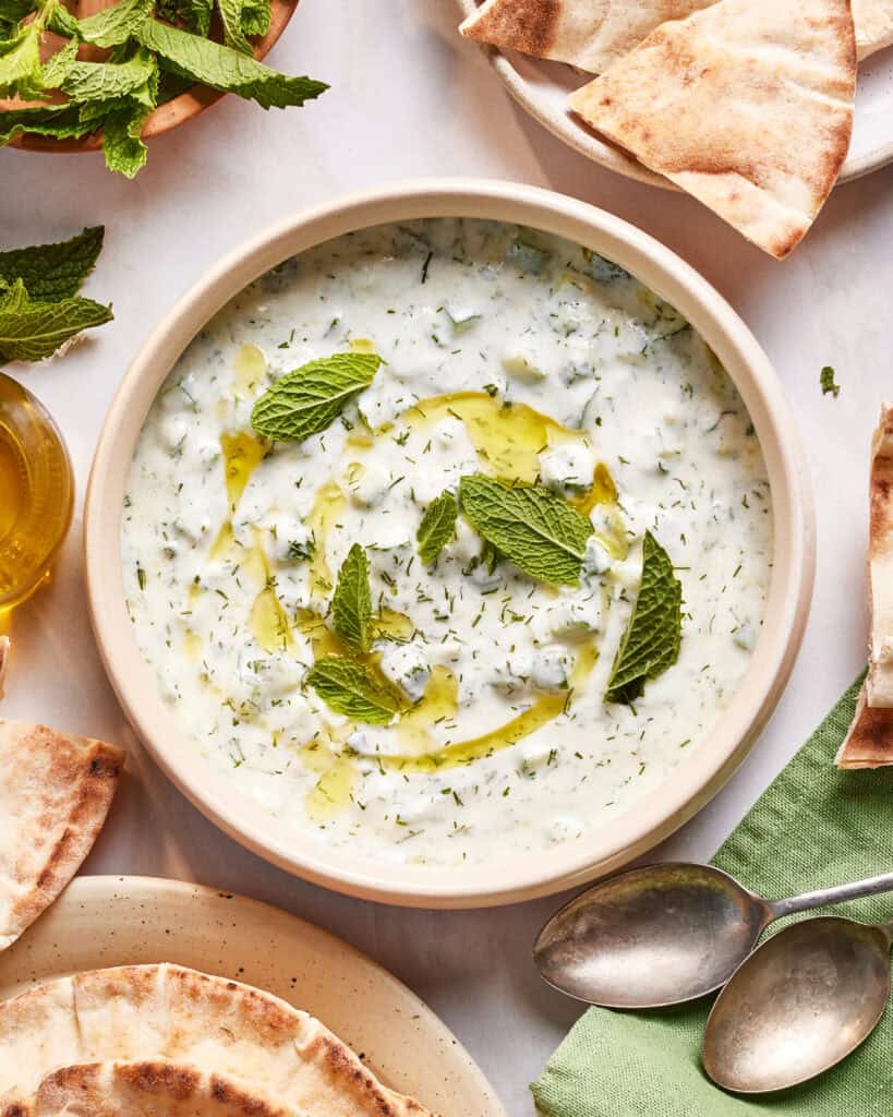 A bowl of tzatziki sauce garnished with fresh mint leaves and olive oil, surrounded by pieces of pita bread, whole mint sprigs, and two metal spoons on a green napkin.