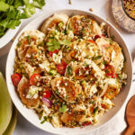 A bowl filled with crispy dumplings, shredded cabbage, cherry tomatoes, peanuts, and herbs. The dish is garnished with a sprinkle of crushed peanuts and cilantro. Additional bowls with peanuts and sauce are visible in the background.