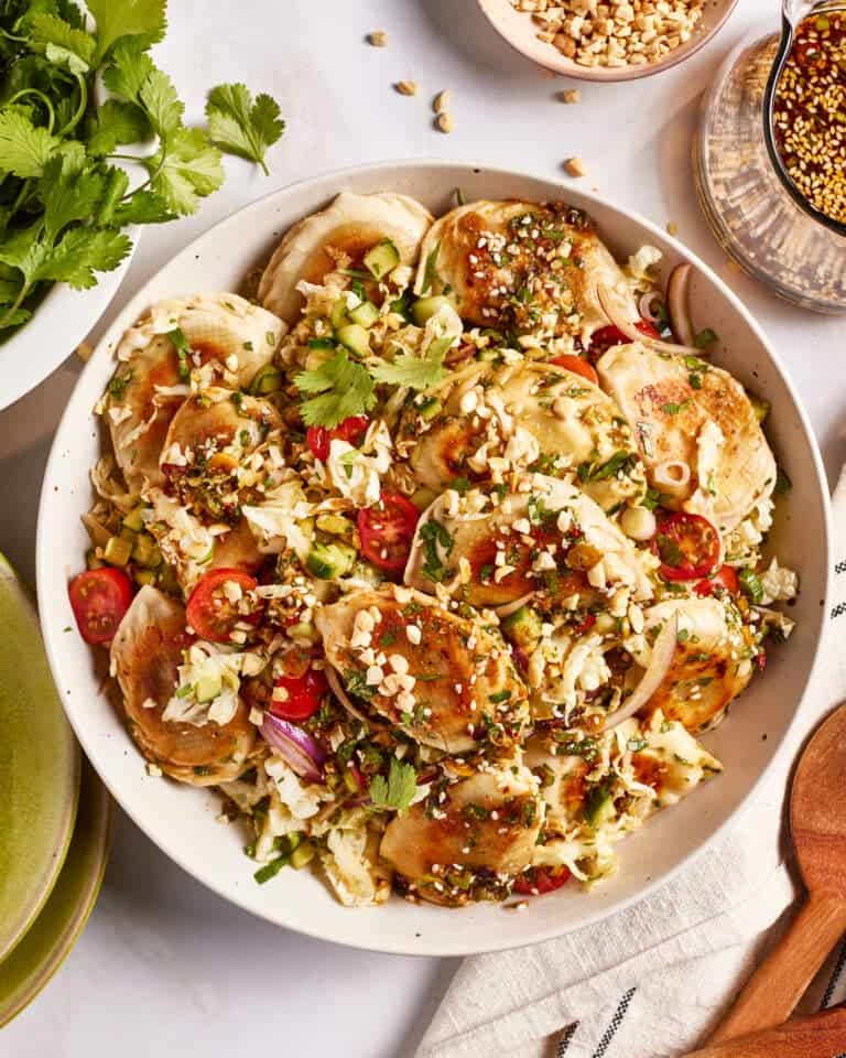 A bowl filled with crispy dumplings, shredded cabbage, cherry tomatoes, peanuts, and herbs. The dish is garnished with a sprinkle of crushed peanuts and cilantro. Additional bowls with peanuts and sauce are visible in the background.