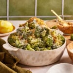 A bowl of roasted broccoli topped with grated cheese and breadcrumbs. The dish is served in a white bowl placed on a table with a green cloth. A lemon, a wooden spoon, and a bowl of breadcrumbs are in the background.