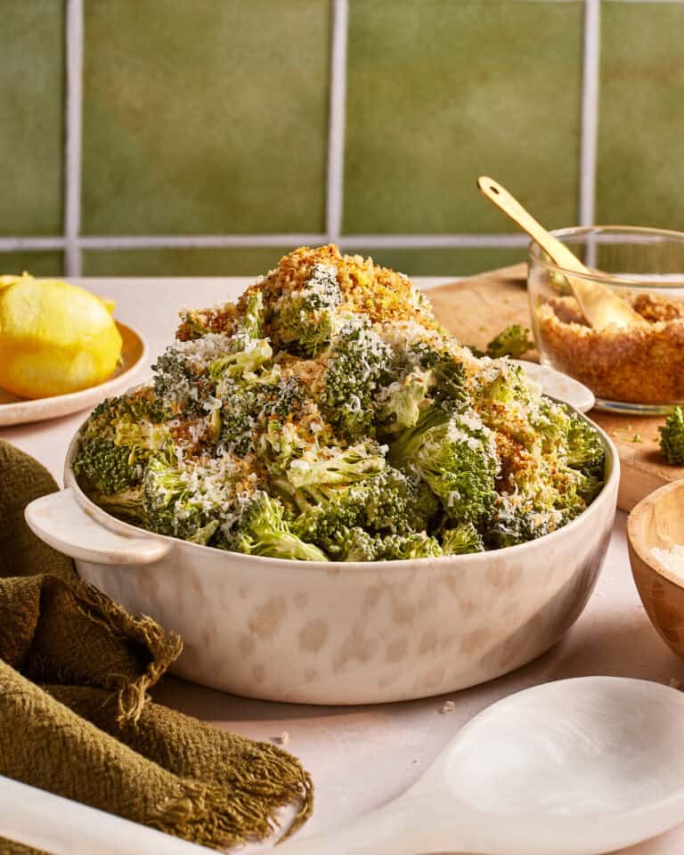 A bowl of roasted broccoli topped with grated cheese and breadcrumbs. The dish is served in a white bowl placed on a table with a green cloth. A lemon, a wooden spoon, and a bowl of breadcrumbs are in the background.