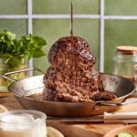A vertical stack of cooked ground meat resembling shawarma is mounted on a skewer in a pan. Surrounding it are fresh mint leaves, a jar of seasoning, sliced cucumbers, white sauce, and bread on a wooden surface with a green tiled backdrop.