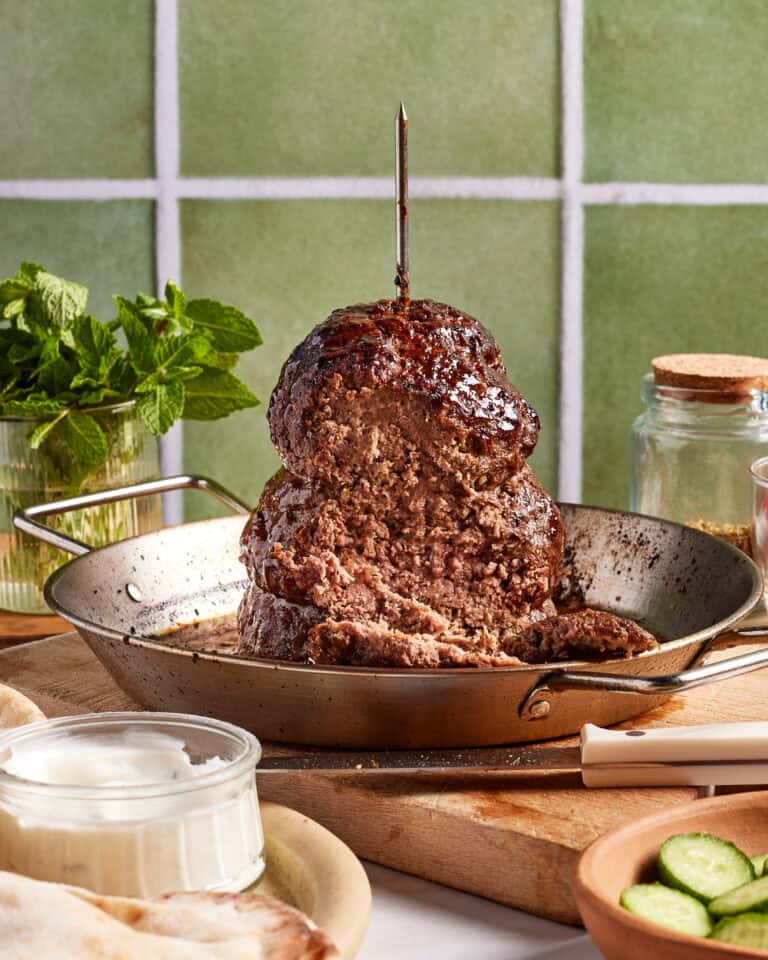 A vertical stack of cooked ground meat resembling shawarma is mounted on a skewer in a pan. Surrounding it are fresh mint leaves, a jar of seasoning, sliced cucumbers, white sauce, and bread on a wooden surface with a green tiled backdrop.