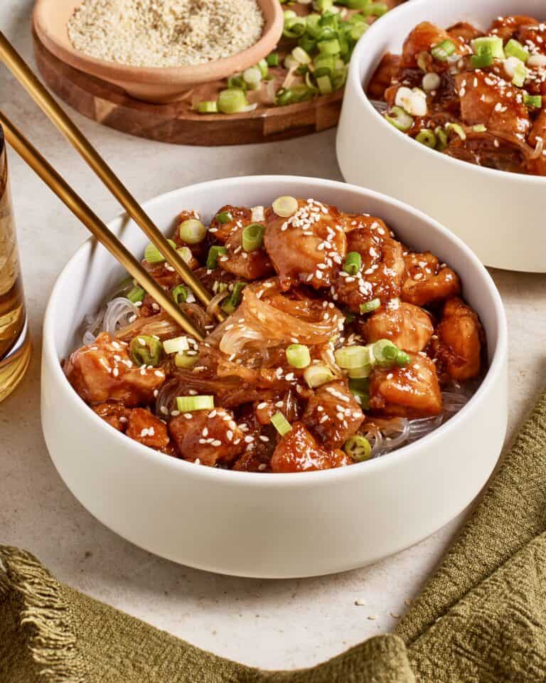 A bowl of noodles topped with glazed pieces of chicken, sliced green onions, and sesame seeds. Chopsticks rest on the bowl. Another similar bowl is partially visible. A small wooden dish with sesame seeds is in the background.