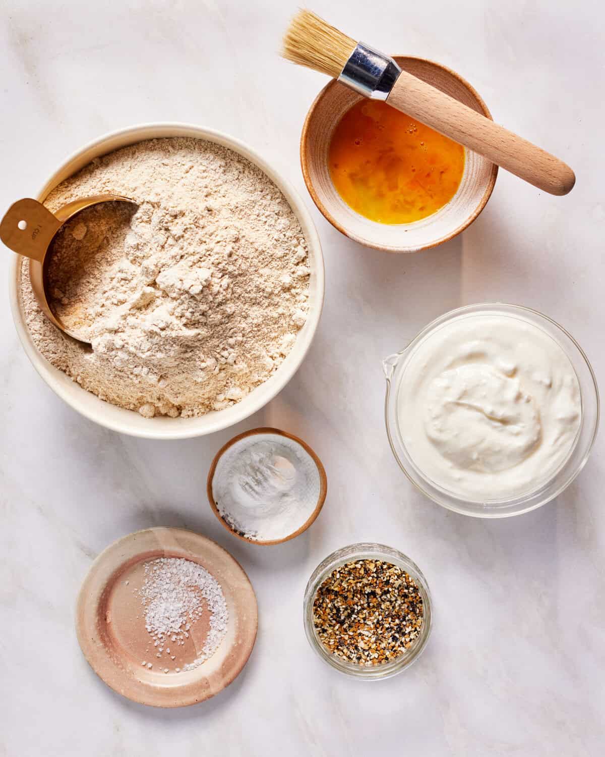 Top view of baking ingredients on a marble surface. Includes a bowl of flour with a wooden scoop, beaten egg in a bowl with a brush, a bowl of yogurt, small dishes of salt, baking powder, and a mix of seeds and spices.