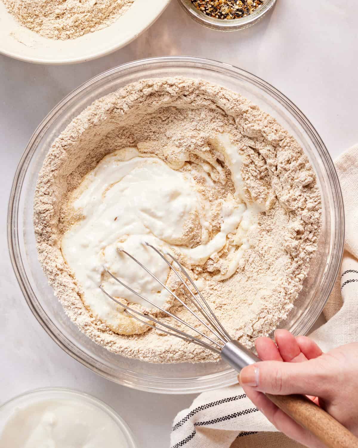 A hand uses a whisk to mix yogurt into a bowl of dry ingredients, likely flour. Various other ingredients are visible around the bowl on a light countertop, suggesting the preparation of a recipe.