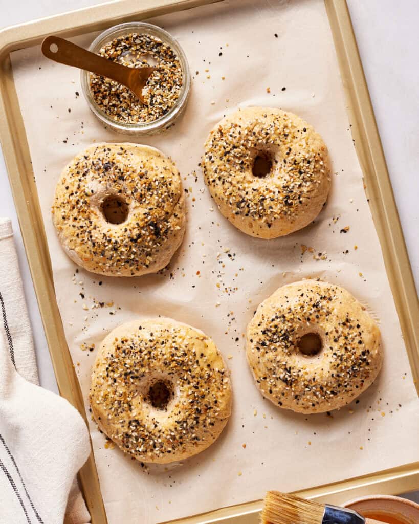 Four unbaked bagels topped with seed mixture on a parchment-lined baking sheet. A small bowl of seeds with a wooden spoon and a basting brush are nearby on a light-colored surface, along with a folded striped cloth.