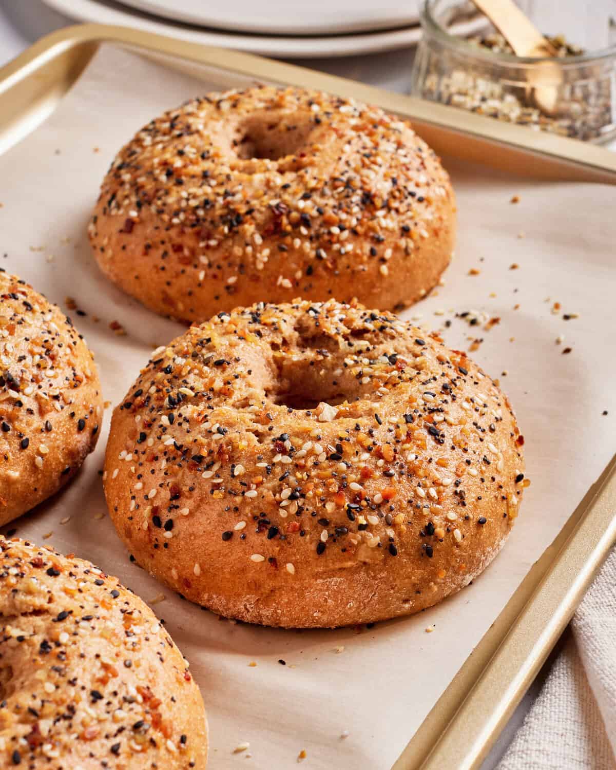 Three freshly baked everything bagels on a parchment-lined baking sheet. The bagels are topped with a variety of seeds and seasonings. In the background, there is a small jar of seasoning and stacked white plates.