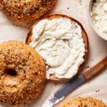 Top view of everything bagels on parchment paper. One bagel is sliced open with creamy spread on it. A knife with more spread lies nearby, and a bowl with the same spread is partially visible in the upper right corner.