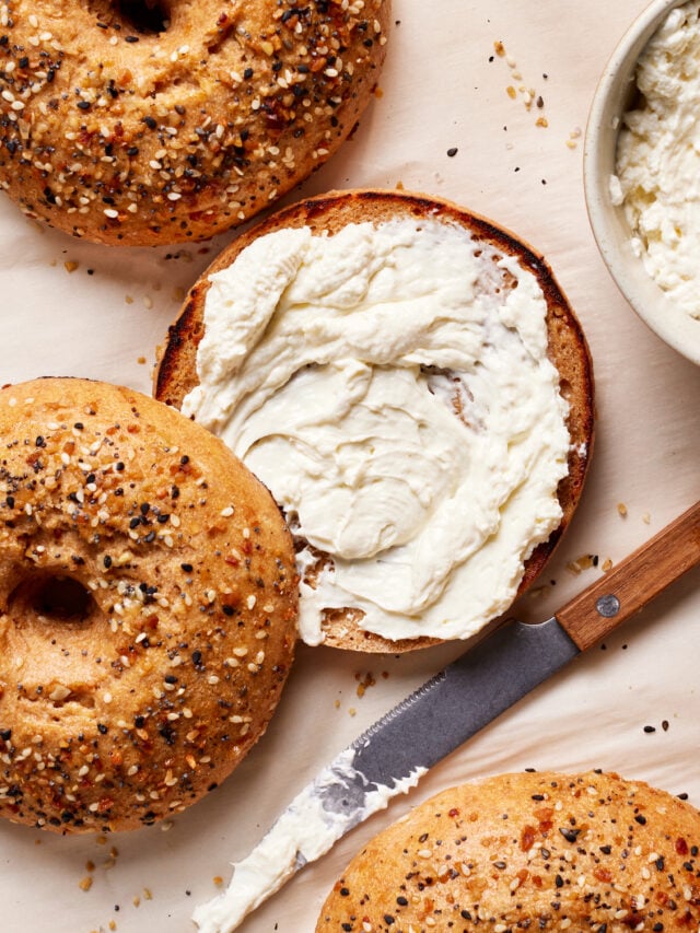 Top view of everything bagels on parchment paper. One bagel is sliced open with creamy spread on it. A knife with more spread lies nearby, and a bowl with the same spread is partially visible in the upper right corner.