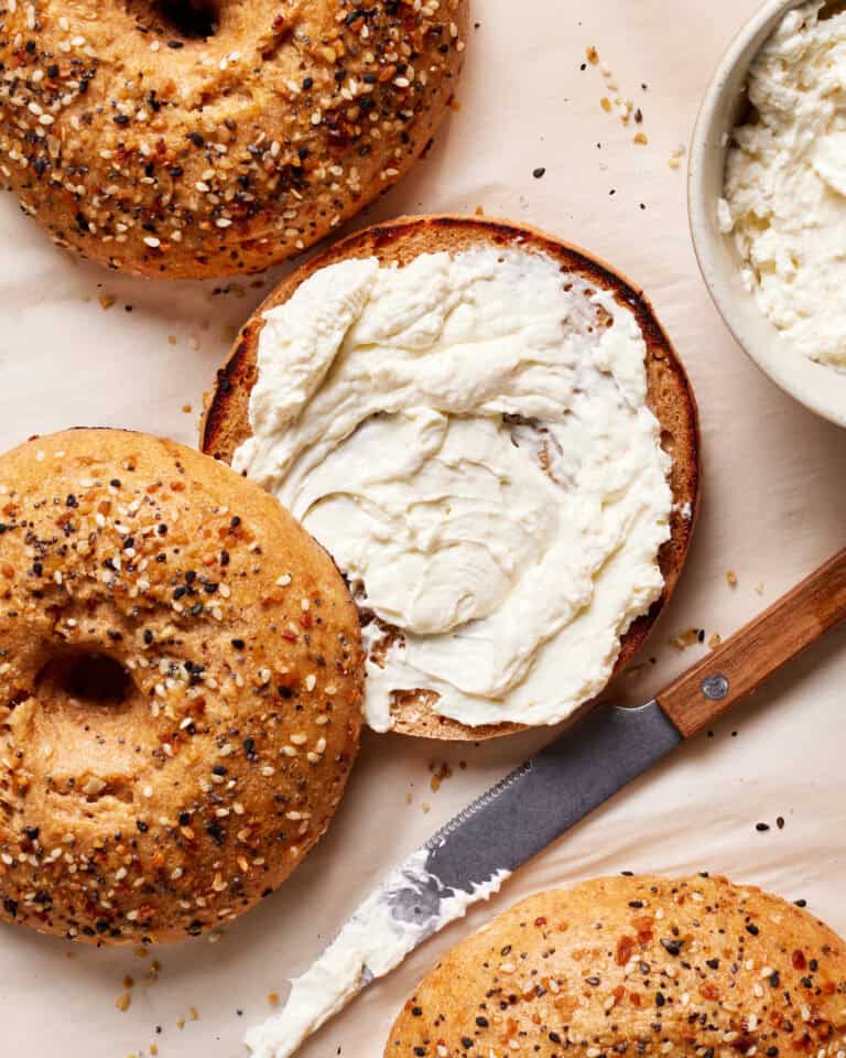 Top view of everything bagels on parchment paper. One bagel is sliced open with creamy spread on it. A knife with more spread lies nearby, and a bowl with the same spread is partially visible in the upper right corner.