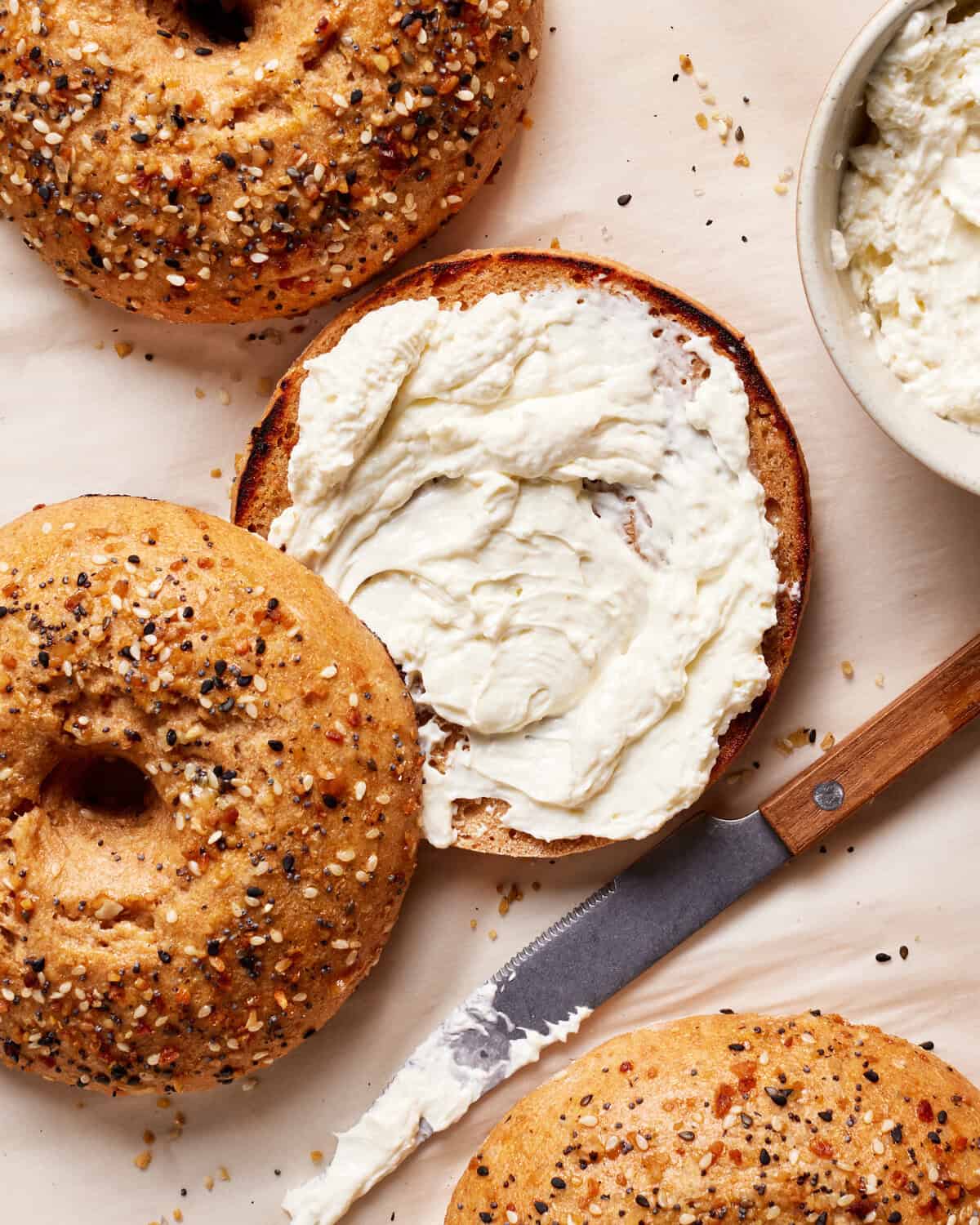 Top view of everything bagels on parchment paper. One bagel is sliced open with creamy spread on it. A knife with more spread lies nearby, and a bowl with the same spread is partially visible in the upper right corner.