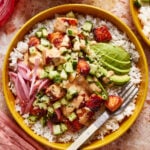 A colorful bowl with white rice, topped with diced cucumber, sauce-covered cubes, sliced avocado, and pickled red onions. A fork rests on the side of the bowl on a pink textured surface.