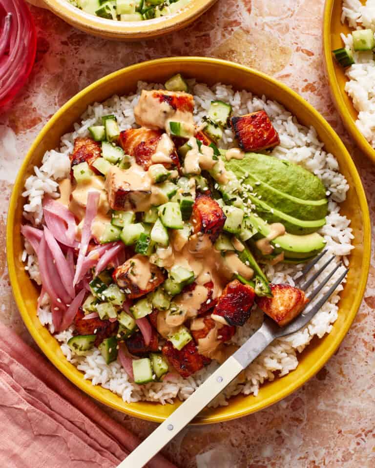 A colorful bowl with white rice, topped with diced cucumber, sauce-covered cubes, sliced avocado, and pickled red onions. A fork rests on the side of the bowl on a pink textured surface.